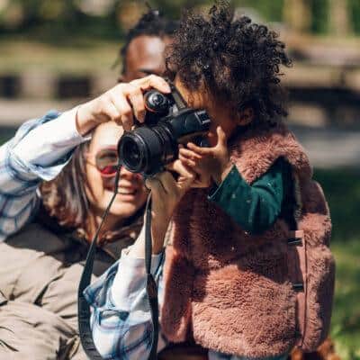 Beautiful multiracial family relaxing in the park during a sunny day and using a digital camera. Photographing memories. African american baby girl holding a camera. Baby girl holding a camera.