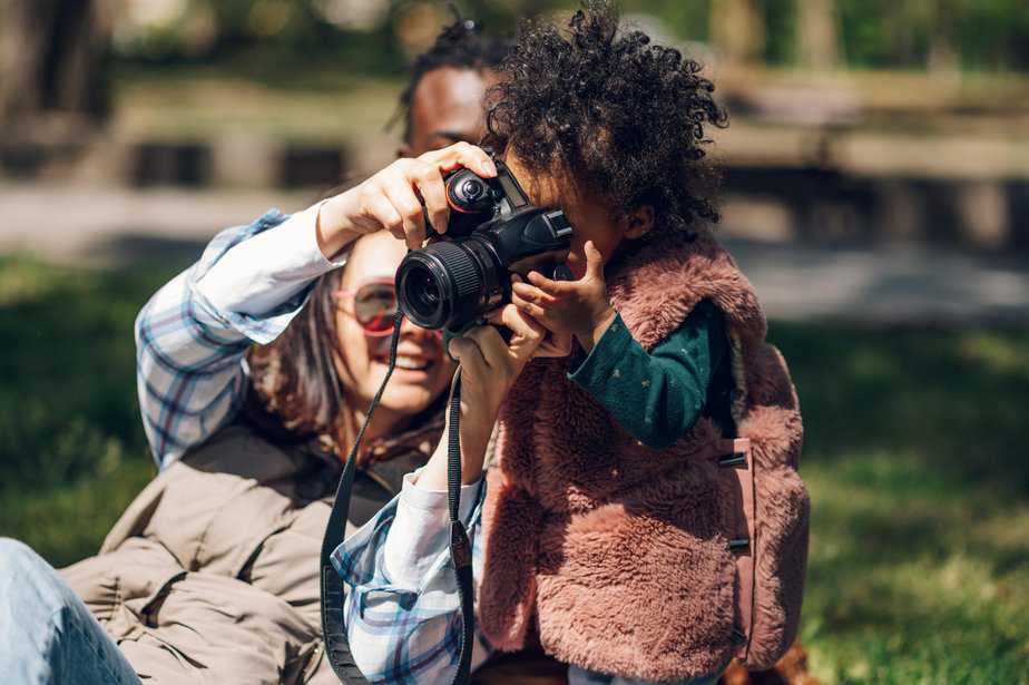 Beautiful multiracial family relaxing in the park during a sunny day and using a digital camera. Photographing memories. African american baby girl holding a camera. Baby girl holding a camera.